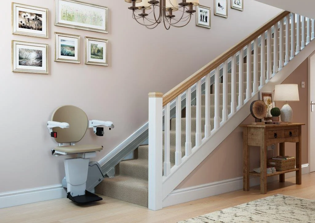 A stairlift with a cream seat parked at the foot of a carpeted staircase in a bright hallway with framed pictures on the wall.